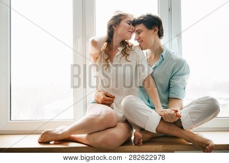 Happy Girl And Man Hugging Near Window In Home. White And Blue Clothes. Valentine Day.