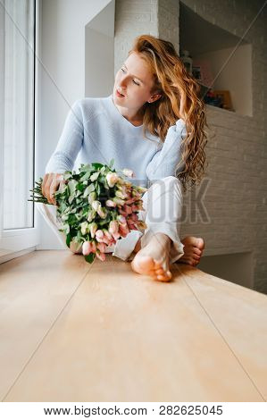 Portrait Of A Young Beautiful Girl With A Bouquet Of Roses Near Window. Curly Flowing Red Hair.
