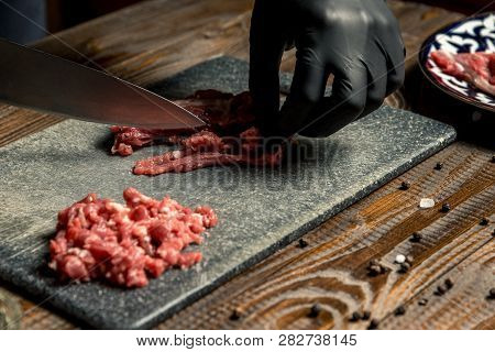 Hands Of The Cook In Gloves Cut The Red Meat On The Board. Nearby Are Tomatoes, Onion, Butter