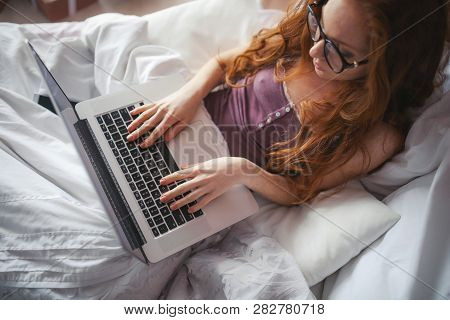 Red-haired Girl Sitting On Bed. Shes Talking By Telephone And Doing Something On Laptop. Shes Lookin