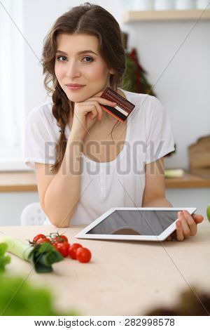 Young  Woman Looking For A New Recipe For  Cooking In A Kitchen. Housewife Is Making Online Shopping