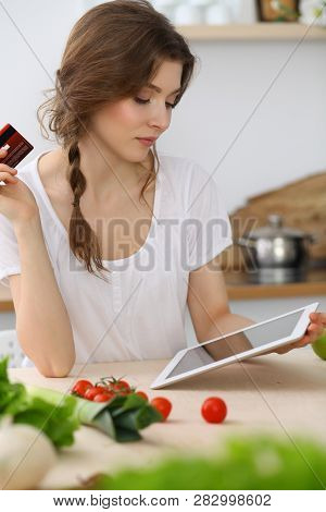 Young  Woman Looking For A New Recipe For  Cooking In A Kitchen. Housewife Is Making Online Shopping