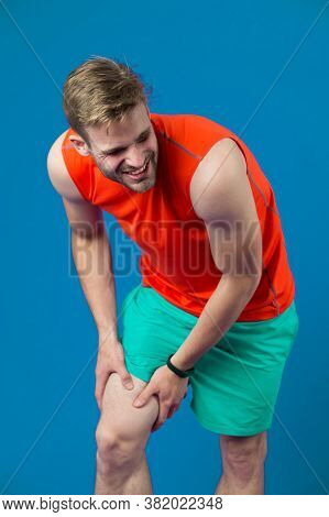 Sportsman With Knee Pain On Violet Background. Man In Orange Vest And Blue Shorts. Health Problem An