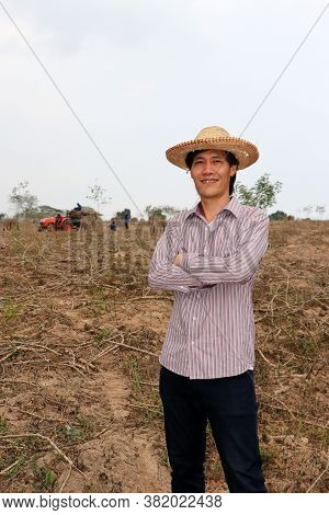 Male Farmer Standing And Hugging Chest In The Cassava Farm With Out Focus Tractor And Worker.