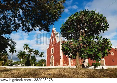 Red Colonial Church Itzimna In A Sunny Park Framed By Trees, Merida, Yucatan, Mexico