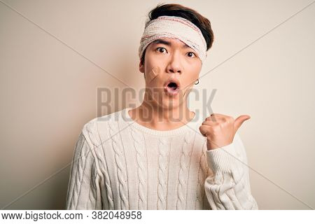 Young handsome chinese man injured for accident wearing bandage and strips on head Surprised pointing with hand finger to the side, open mouth amazed expression.