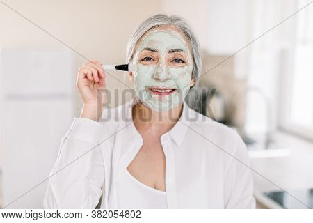 Pretty Elderly Caucasian Woman With Grai Hair, Posing To Camera In Modern Home Kitchen While Applyin