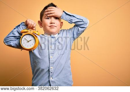 Young little boy kid holding classic bell alarm clock over isolated yellow background stressed with hand on head, shocked with shame and surprise face, angry and frustrated. Fear and upset for mistake