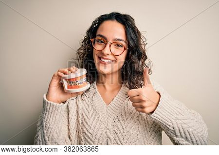 Young beautiful woman with curly hair holding plastic teeth with dental baces happy with big smile doing ok sign, thumb up with fingers, excellent sign
