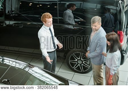 Middle aged consumers in smart casualwear listening to young sales manager showing new model ofo car and describing its characteristics