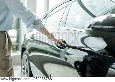 Hand of young elegant man in shirt and trousers standing by luxurious black car and holding by handle while going to open one of doors