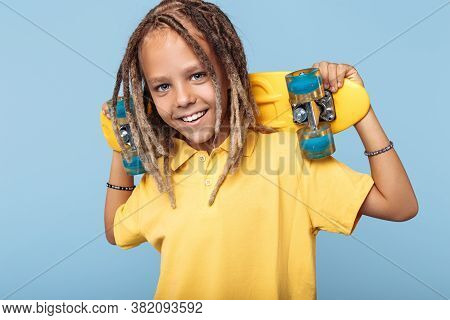 Happy Little Boy With African Dreads Holding Skateboard On Shoulders And Smile Over Blue Background.