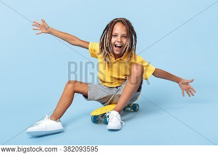 Cheerful Little Boy With African Dreads Sitting On Skateboard Over Blue Background.
