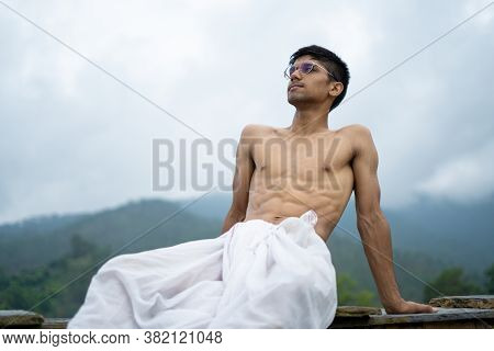 Young Shredded Man Sitting Wearing A Dhoti And Glasses With Blurred Mountains In The Background.