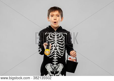 halloween, holiday and trick-or-treating concept - excited boy in black costume of skeleton with candies and flashlight over grey background