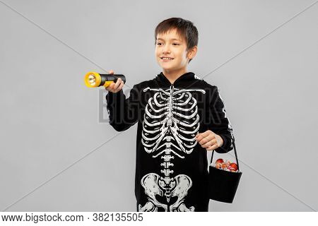 halloween, holiday and trick-or-treating concept - smiling boy in black costume of skeleton with candies and flashlight over grey background