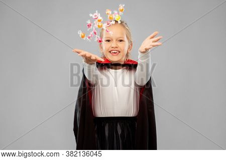 halloween, holiday and childhood concept - happy smiling girl in black dracula cape or costume with candies over grey background