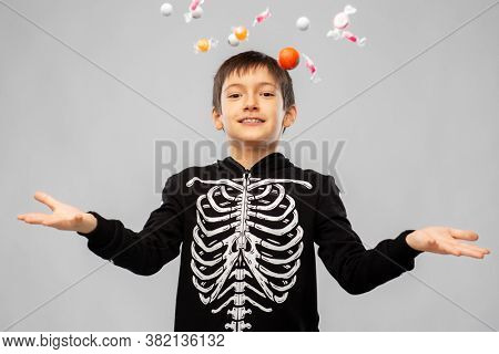 halloween, holiday and childhood concept - smiling boy in black costume of skeleton with candies over grey background