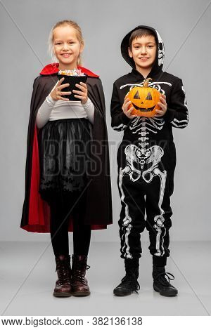 halloween, holiday and trick-or-treating concept - happy smiling boy and girl in costumes of dracula and skeleton with candies and jack-o-lantern pumpkin over grey background
