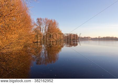 Pond Surrounded By Trees With Fallen Leaves In Windless Calm Weather Against The Clear Sky In Mornin