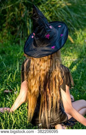 Causcasian Blond Girl With Long Hair In Halloween Witch Costume Sitting On A Green Grass.