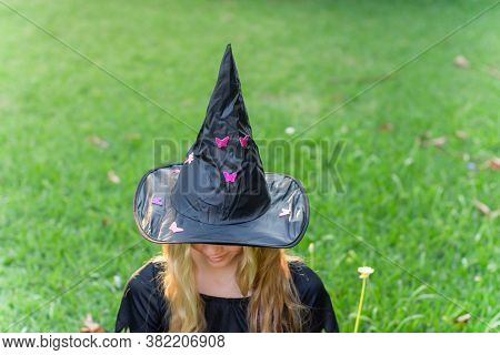Causcasian Blond Girl With Long Hair In Halloween Witch Costume Sitting On A Green Grass.