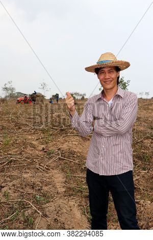 Male Farmer Standing And Thumbs Up In The Cassava Farm With Out Focus Tractor And Worker.