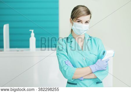 Female Doctor Or Nurse In Protective Mask At Hospital Holds Pyrometer In Hand. Safety Measures Again