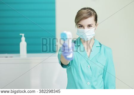 Female Doctor Or Nurse In Protective Mask At Hospital Holds Pyrometer In Hand. Safety Measures Again