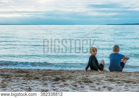 Two Young Brothers Sitting On A Beach In St Heliers, Auckland, New Zealand. The Older Brother Is Loo