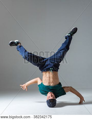 Stylish Guy Dancer Dancing On Head Breakdance In Studio Isolated On Gray Background. Breakdance Less