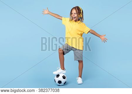 Cheerful Little Boy With African With Headphones Holding One Leg On Soccer Ball Over Blue Background