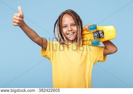 Positive Little Boy With African Dreads Holding Skateboard On Shoulders And Showing Thumb Up Over Bl