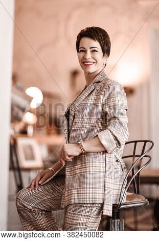 Positive Short Haired Brunette Woman In Elegant Business Plaid Pantsuit With Rolled Up Sleeves Sits 
