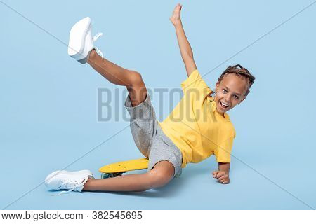 Positive Little Boy With African Dreads Sitting On Skateboard Having Fun Over Blue Background.