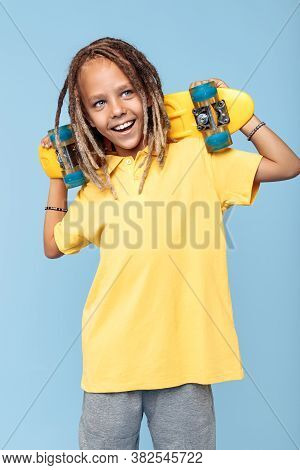 Friendly Little Boy With African Dreads Holding Skateboard On Shoulders And Smile Over Blue Backgrou