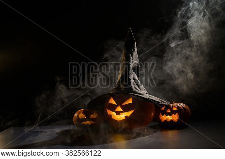 Three Jack O Lanterns Glow In The Dark Amidst The Fog. Halloween Pumpkin In A Witch Hat.