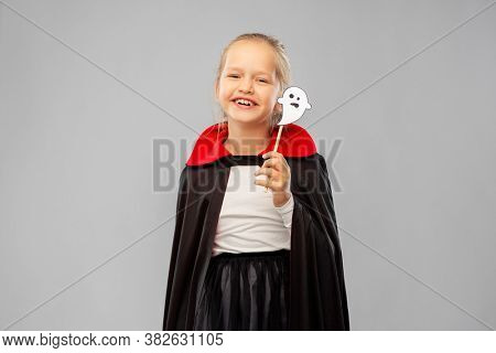 halloween party, holiday and childhood concept - happy smiling girl in black dracula costume with cape and ghost over grey background