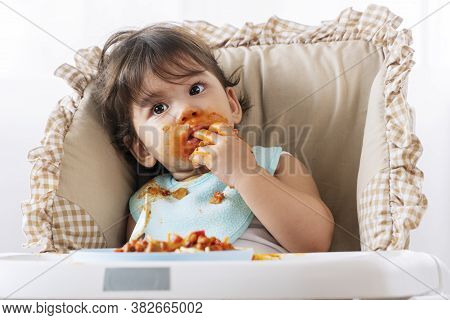 Adorable Little Funny Girl Of 12 Months Eating Spaghetti With Spoon While Sitting In High-powered Ch