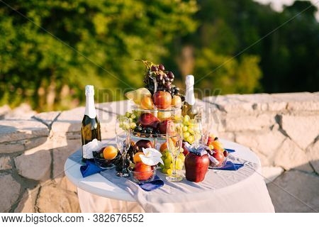 Round Table Covered With White Tablecloth With Full Bottles Of Champagne, Glasses And Fruits In Natu