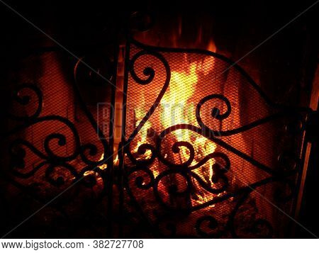 Fire In The Fireplace Behind A Beautiful Wrought Iron Grate With Curls. A Fire Is Burning In The Fir