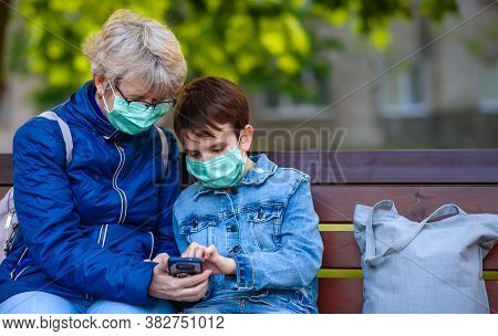 Boy 10 Years Old Is Sitting In Park With Grandmother In Medical Mask With A Smartphone In Hands. He 