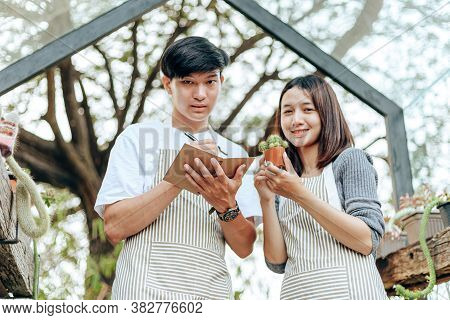 Women Hold Cactus And A Man Writing Note In A Book. Love Couple Enjoy Hobby With Darden Cactus.