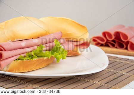 Mortadella Sandwich And Lettuce On A White Plate On A Mat And Slices Of Mortadella Next To It On A C