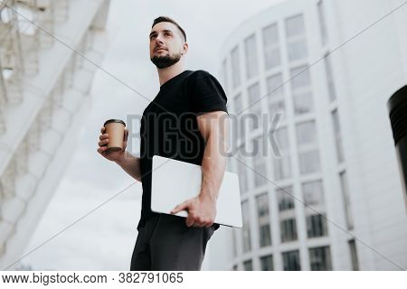 Young Handsome Fit Man Walking Down The Street, Carrying His Laptop While Drinking Coffee On The Go,