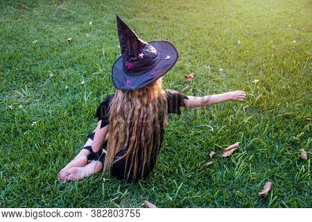 Causcasian Blond Girl With Long Hair In Halloween Witch Costume Sitting On A Green Grass