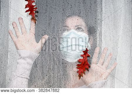 Woman In Medical Mask Indoors Looking Through Glass Window In Raindrops Looks At Falling Autumn Leav