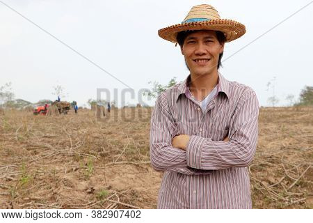 Male Farmer Standing And Hugging Chest In The Cassava Farm With Out Focus Tractor And Worker.