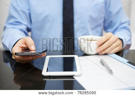 Businessman Is Using Smart Phone And Reading Email On Tablet Pc And Holding Cup Of Coffee With Docum