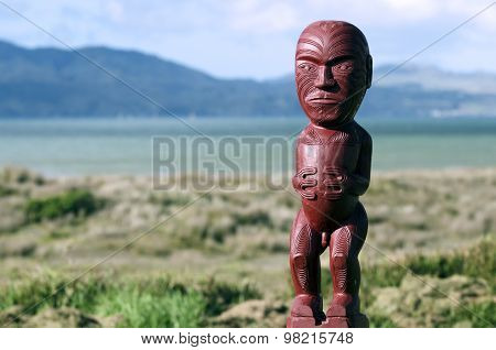 Red wood carving of Maori figurine with New Zealand landscape in the background.